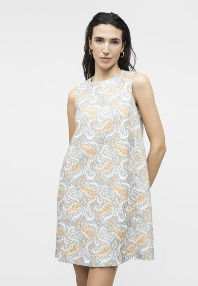 Γυναίκα με μακριά σκούρα μαλλιά, φορεμένη με αμάνικο φόρεμα μήκους μέχρι τα γόνατα, με μπλε και μπεζ λουλουδάτο μοτίβο παϊέτας σε ανοιχτό φόντο.