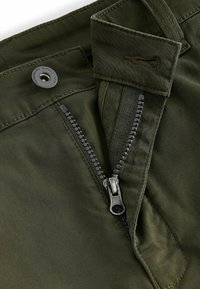 Pantaloni de culoare verde măsliniu cu fermoar cu dinți negri, închidere cu nasture și cusături detaliate de-a lungul taliei și în zona buzunarelor.