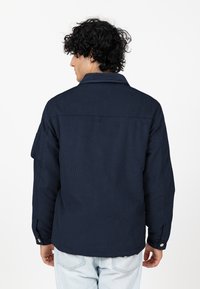 Navyblauwe corduroy jas met een puntige kraag, knopenmanchetten en een patchzak op de linkermouw; een gladde textuur en een getailleerd ontwerp.