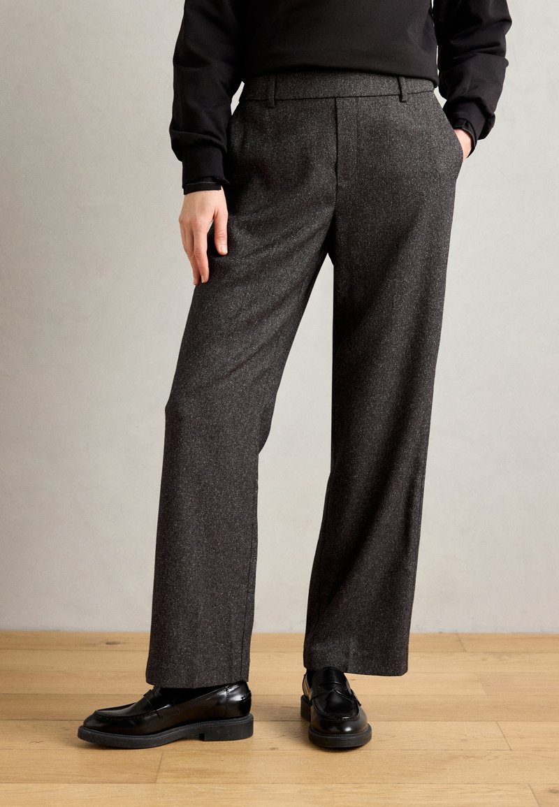 Personne portant un pantalon large gris foncé avec des mocassins noirs, debout sur un sol en bois devant un fond uni.