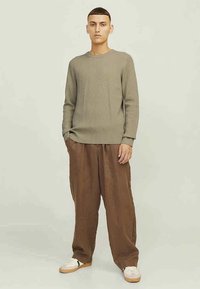 Olivno zelena rebrasta majica s dolgimi rokavi in krožnim izrezom, v kombinaciji z ohlapnimi rjavimi hlačami in belimi supergami z zelenimi poudarki.