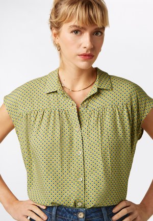 Zielona bluzka w wzory z krótkimi rękawami, kołnierzem i guzikami. Wykonana z fakturowanej tkaniny z geometrycznym wzorem w żółtym i niebieskim kolorze.