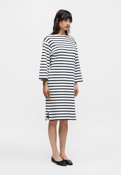 Γυναίκα που φοράει λευκό φόρεμα μέχρι το γόνατο με μαύρες οριζόντιες ρίγες και μανίκια 3/4, σε συνδυασμό με μαύρα επίπεδα παπούτσια, στέκεται σε λευκό φόντο.