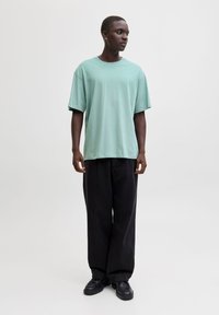 Jasnozielona, krótka koszulka z miękkiego materiału, zestawiona z czarnymi, luźnymi spodniami i czarnym obuwiem. Neutralne tło.