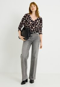 Mujer con cabello rojo que lleva una blusa de estampado de leopardo, jeans grises de talle alto, botas negras y sostiene un bolso negro.
