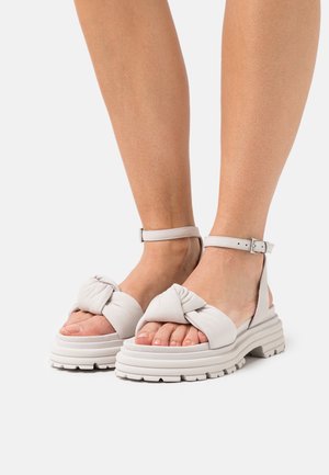 Light gray platform sandals with knotted fabric straps and ankle buckle. Thick, textured soles with a ridged pattern for traction.
