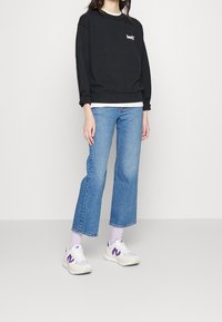 Sweatshirt noir avec logo blanc, superposé sur une chemise blanche. Jean bleu à jambes larges, baskets blanches et violettes, stylisé avec des chaussettes montant à la cheville.