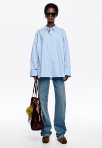 Lichtblauwe oversized shirt, met knopen; gecombineerd met flare jeans. Accessoires omvatten een bruine leren tas met een gele vacht details.