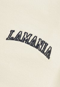 Kremo spalvos audinys su tamsiai mėlynu siuvinėtu tekstu "LAMANIA" išlenkto dizaino. Tekstūra yra lygi ir minkšta, matomi aiškūs siuvimo detalės.