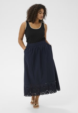 Woman with curly hair wearing a black sleeveless top and navy blue midi skirt with floral cutout hem, standing with hands in pockets.