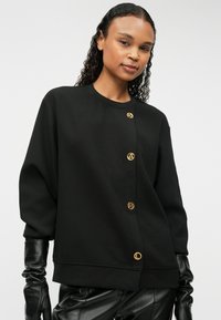 Μαύρο φούτερ μπουφάν με στρογγυλή λαιμόκοψη, διακοσμημένο με τέσσερα χρυσά κουμπιά λογότυπου μπροστά και μανίκια τύπου μπαλονιού.