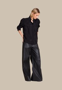 Svart, kortermet skjorte med rynkede skuldre, kombinert med vide, høyt midje svarte skinnbukser med både teksturerte og glatte overflater.