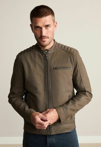 Braune Lederjacke mit Stehkragen, senkrechtem Reißverschluss und gepolsterten Schulterakzenten. Sanfte Textur und taillierte Passform.