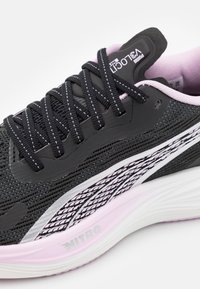 Gros plan sur une chaussure de course noire et rose avec une semelle intermédiaire à motifs, de grosses lacets noirs et un col rembourré. La chaussure arbore les marques "V3 Loci" et "Nitro".