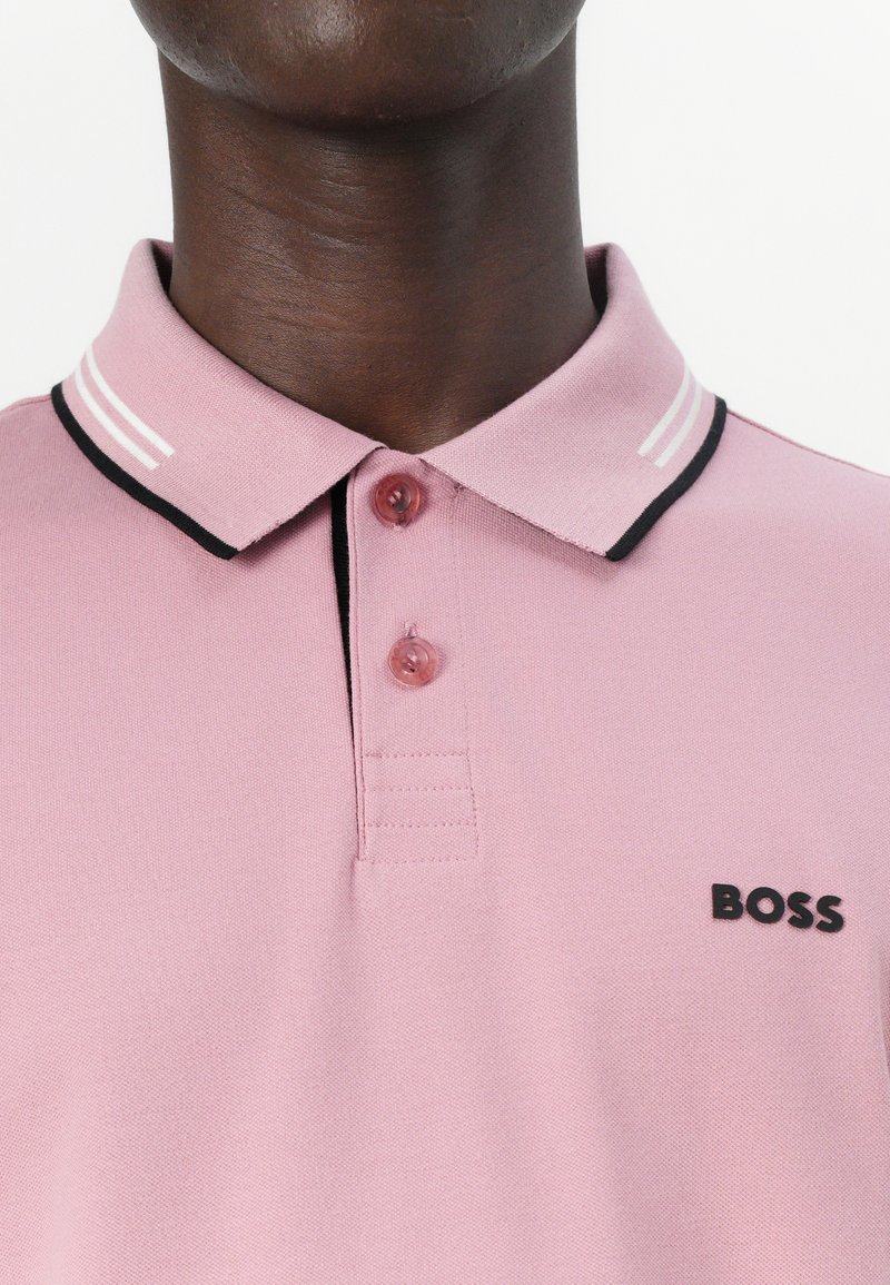 Camisa polo rosa claro con tela texturizada, detalles en negro en el cuello y un logo negro de "BOSS" en el lado izquierdo del pecho. Botonera de tres botones.