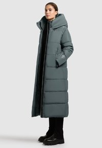 Cappotto lungo e voluminoso di colore grigio medio, con un grande cappuccio, chiusura con zip, tasche laterali e polsini a coste, abbinato a stivaletti neri con tacco alto.