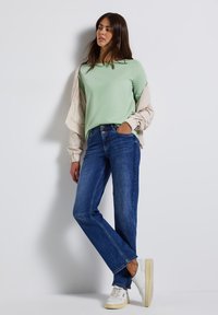 Chemise verte clair à manches longues superposée avec une veste bomber beige, accompagnée de jeans en denim bleu et de baskets blanches, se tenant devant un fond uni.