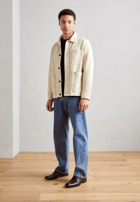 Chaqueta beige clara con botones y bolsillos de parche, camisa negra debajo, jeans de mezclilla azul y zapatos de vestir marrones con un acabado suave.