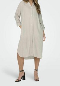 Abito camicia in lino beige chiaro con maniche lunghe, bottoni frontali, spacco laterale e vestibilità ampia, abbinato a sandali neri con tacco a blocco.