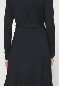 Vue de dos d'une personne portant une longue robe noire à manches longues et une ceinture cintrée à la taille.