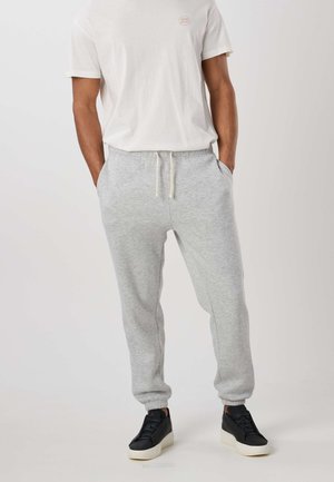 Homme portant un pantalon de survêtement gris clair à cordon, un T-shirt blanc et des baskets noires avec des semelles blanches, debout les mains dans les poches.