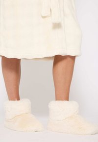 Zapatillas de crema difusas con puño plegable, textura suave y forma cónica, que brindan calidez y comodidad durante su uso en interiores.