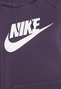 Fialová mikina s kapucí z měkké látky, na které je velké bílé logo Nike a přední kapsa. Obsahuje stahovací šňůrku s černými konci.
