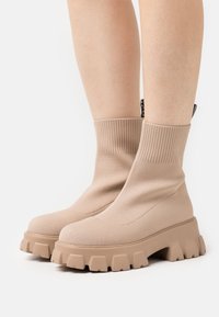 Bottes chevilles beiges avec une tige en tricot côtelé, semelle en caoutchouc épaisse et tissu texturé. Présente une étiquette logo à l'arrière pour le détail.