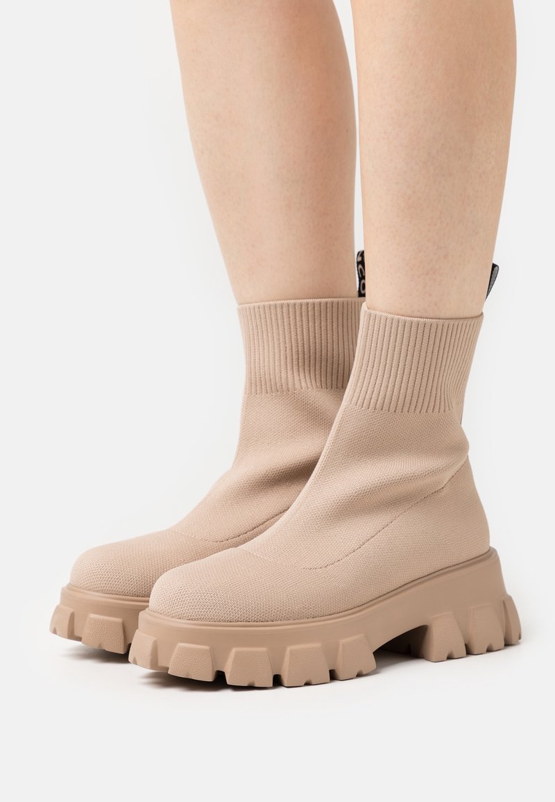 Bottes chevilles beiges avec une tige en tricot côtelé, semelle en caoutchouc épaisse et tissu texturé. Présente une étiquette logo à l'arrière pour le détail.