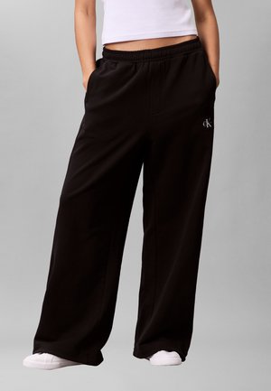 Person wearing black wide-leg pants with elastic waist and small logo on thigh, paired with white sneakers and cropped white top.