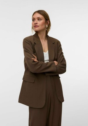 Vrouw met lichtbruin haar draagt oversized bruinen pak en witte top, staat met gekruiste armen, kijkt opzij tegen een witte achtergrond.