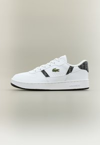 Λευκό αθλητικό παπούτσι Lacoste με μαύρες γεωμετρικές λεπτομέρειες, πράσινο λογότυπο κροκοδείλου στο πλάι και λευκή σόλα με την εγχάρακτη λέξη "Lacoste" στην φτέρνα.