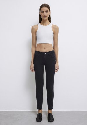 Woman standing against white wall wearing white sleeveless crop top, black skinny jeans, and black loafers, facing forward with hands relaxed.