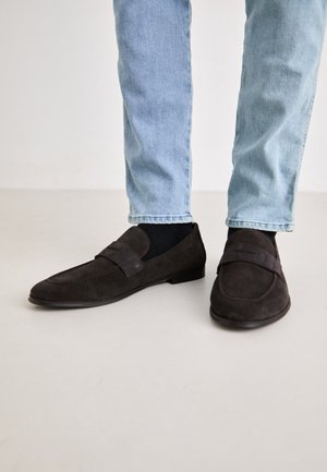 Donkerbruine suède instappers met een platte zool en een brede band over de bovenkant. G gedragen met lichtblauwe spijkerbroek en zwarte sokken.