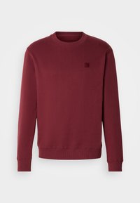 Sweat-shirt à manches longues col rond en bordeaux profond avec poignets et ourlet côtelés, avec un petit logo carré cousu sur la poitrine gauche.