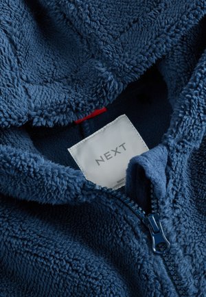 Laivastonsininen fleece-huppari, jossa on teksturoitu, pörröinen materiaali. Siinä on vetoketjusuljenta ja näkyvä valmistajan etiketti, jossa lukee "NEXT".