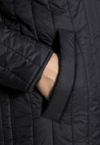 Chaqueta aislante negra con un diseño acolchado, bolsillo lateral y forro interior texturizado. El material parece ligero y brillante.