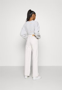Jeans de perna larga de cintura alta em bege claro, com um bolso nas costas e uma etiqueta de couro bege. O modelo usa um sweatshirt oversized cinza e ténis brancos.