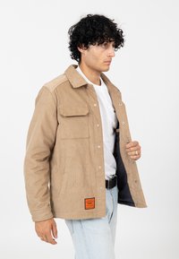 Beżowa kurtka sztruksowa z kołnierzykiem, dwiema przednimi kieszeniami, srebrnymi zatrzaskami i pomarańczową metką na końcu. Noszona na białym t-shircie.