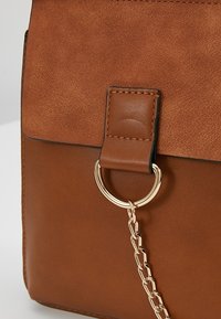 Sac en cuir marron avec rabat en daim, doté d'une boucle en cuir cousue attachée à un anneau métallique doré et un détail en chaîne.