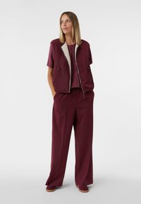 Gilet bordeaux con foderatura in pelliccia sopra un top coordinato e pantaloni a gamba larga. L'insieme presenta una vestibilità rilassata e sottili accenti testurizzati.