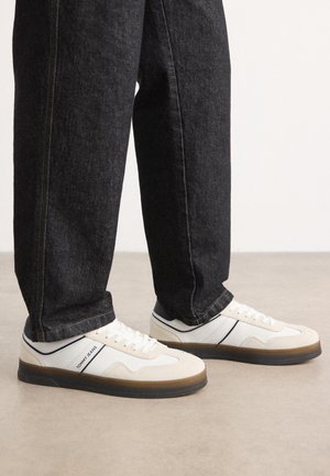 Weiße und hellgraue Sneakers mit einem strukturierten Obermaterial aus Wildleder und Leder, brauner Gummisohle und navyblauem Akzentstreifen. Getragen mit dunklen Jeans.