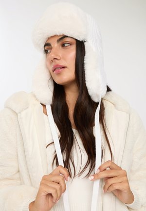 Cappello trapper in pelliccia sintetica bianca con design lavorato a maglia, orecchiette staccabili e lacci bianchi. Texture morbida, materiale spesso e copertura completa.