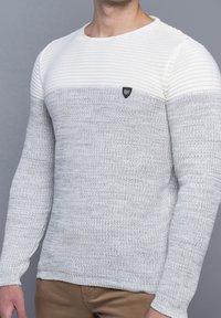 Pull en maille gris clair et crème avec texture côtelée, manches longues et un petit logo noir sur la poitrine. Encolure ronde classique.