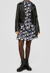 Kvinna iklädd en svart läderjacka över en blå, svart och vit blommig klänning, svarta strumpbyxor och svarta platåstövlar med snörning.