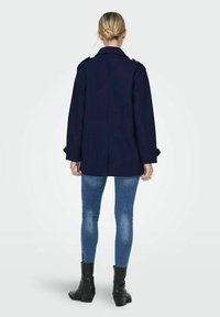 Manteau bleu marine au design structuré, doté d'un col haut et d'accents de boutons, associé à un jean slim et des bottines.