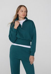 Turkusowy rozpinany sweatshirt z wysokim kołnierzem i elastycznym pasem, w komplecie z dopasowanymi spodniami. Sweatshirt posiada biały wewnętrzny materiał.