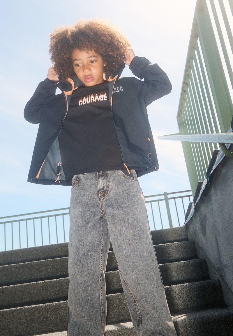 Giacca nera con zip e dettagli arancioni, t-shirt nera con il testo "COURAGE" e jeans a gamba larga grigio chiaro su una persona su scale di cemento.