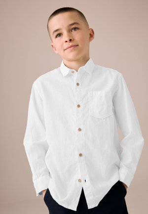 Dreng med kort hår iført en hvid skjorte med knapper og mørke bukser, stående med hænderne i lommerne mod en neutral baggrund.