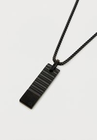 Collier avec pendentif noir de forme rectangulaire, présentant une surface rayée texturée et une base lisse, attaché à une chaîne.